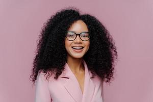 Close up portrait of happy ethnic young woman with crisp hair, looks through optical glasses, wears formal outfit, being in good mood, comes on work, happy get praise from boss, models indoor photo