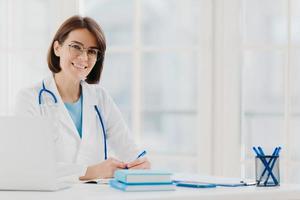 Professional woman doctor writes down notes, poses at desktop in office with laptop, wears white coat, spectacles and phonendoscope around neck, looks through medical documents. Healthcare concept photo