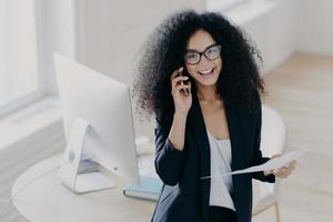 Cheerful female business worker calls partner to discuss details of statrup project, holds paper documents, wears optical glasses and elegant suit, gets necessary information, dressed elegantly photo