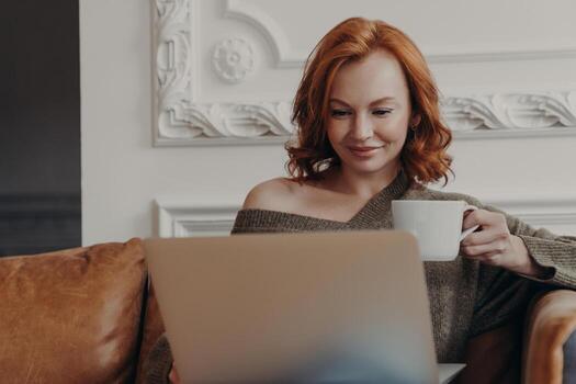 Beautiful redhead European woman concentrated in laptop computer, ponders on completing working tasks, drinks aromatic coffee, sits on comfortable sofa, checks database, analyzes information photo