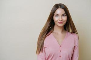 Horizontal portrait of charming European woman smiles gently at camera, has long straight hair, wears casual rosy sweater, going to have outdoor walk, stands against beige studio background. photo