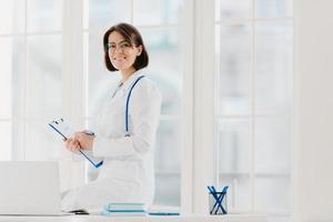 Skilled female physician writes in clipboard, poses at workplace with laptop computer. Professional family doctor ready to give you piece of advice to care about your health, wears medical uniform. photo