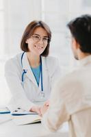 Medical trust concept. Vertical image of female doctor reassures her male patient, hold hand and talk calmly about illness cure, gives professional consultation and support, pose at doctors office photo