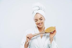 Pretty smiling woman concentrated down, holds bath brush, takes shower, cares about hygiene, wears towel on head, white soft robe, poses against white background. Women and cleanliness concept photo