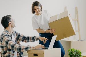 un joven feliz apunta a una computadora portátil, busca un nuevo diseño para una mujer plana y ocupada que lleva cajas, compra una nueva habitación, busca decoraciones para el hogar, se conecta a internet inalámbrico. foto