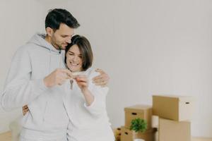 Delighted woman and man home owners enjoy buying new flat, hold keys fromm their own apartment, pose against white wall with unpacked cardboad boxes. Family mortgage, ownership, real estate. photo