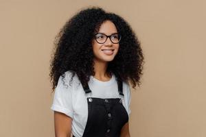 Pleased dreamy woman with curly hairstyle, looks through transparent glasses, dressed in casual white t shirt and black dungarees, poses against brown background, has good mood. People and emotions photo