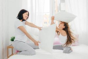 Indoor shot of pleasant looking European female has joy together with her small daughter, have pillow fight in bedroom, pose in cozy spacious light room, being in high spirit. People and fun concept photo