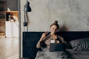 Lovely joyful young girl lying in bed with cup of coffee and having video call with friends online on laptop photo