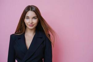 Elegante y exitosa empleada morena europea vestida con traje formal negro, lista para comenzar su propio negocio, se encuentra confiada en el interior, aislada en un fondo rosa. gente, profesión, ocupación foto