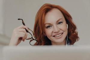 Close up image of ginger positive European woman smiles gently, enjoys online communication, watches tutorial webinar, gets new skills, sits in front of laptop computer, holds transparent glasses photo