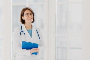 Experienced woman pediatrician stands with clipboard in cabinet, wears white medical coat with stethoscope, gives excellent medical treatment, has happy expression, ready to give consultancy photo