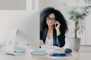 Photo of satisfied Afro American woman calls via smartphone, makes notes in notepad, involved in working process, sits at desk with computer, wears spectacles, formal clothes, has phone communication