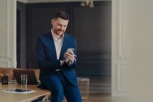 hombre de negocios guapo feliz mirando el teléfono móvil mientras está sentado en el escritorio de la oficina foto