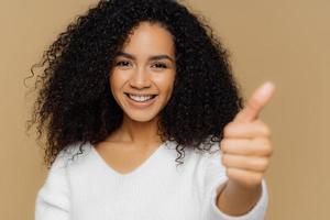 Headshot of dark skinned healthy curly young woman shows thumb up at camera, has pleasant smile, gives approval or likes idea, wears white jumper, isolated on brown studio background. Its okay photo