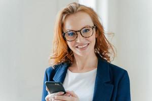 Close up shot of pretty redhead female directror has charming smile, dressed in elegant clothes, holds mobile phone, checks text message, surfs social networks, connected to wireless internet. photo