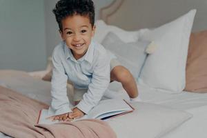 Adirable small afro american smiling child in light blue shirt playing on parents bed photo