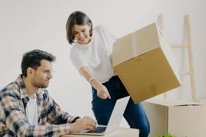 Indoor shot of happy family couple look attentively at laptop computer, search good moving company, carry personal belongings in big cardboard boxes, woman points into display of modern gadget