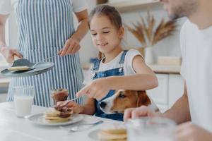 niño pequeño, su padre y su perro se sientan juntos en la mesa de la cocina, comen panqueques recién preparados, la madre en delantal se para cerca de la sartén. postre sabroso apetitoso familiar en la cocina. cocina, concepto de nutrición foto