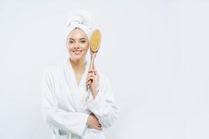 shof horizontal de encantadora mujer de belleza tiene una amplia sonrisa, dientes blancos perfectos, vestido con bata de baño, sostiene un cepillo de masaje anticelulítico, posa contra el fondo blanco, espacio en blanco para su promoción foto