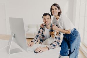 Image of happy brunette smiling woman leans at husbands shoulders who keyboards on computer, surf internet and search new apartment to buy on website, pose in spacious room with domestic dog photo