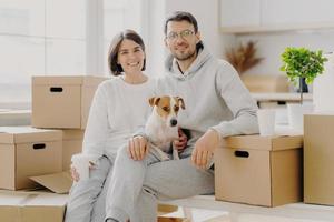 Positive woman and man pose near stack of cardboard boxes, pose for making portrait with dog, relocate in new living place, wear white casual clothes, pose in spacious kitchen with big windows photo