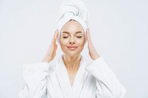 Gorgeous spa woman stands with closed eyes, keeps hands on towel wrapped on head, dressed in white bath robe, has healthy skin, natural makeup, well groomed complexion, poses indoor. Beauty treatment photo