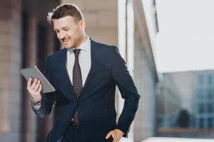 Elegant attractive male executive manager holds modern digital tablet, reads financial news on web page, rejoices having success poses outdoor, has spare time during dinner break. Job and technology photo