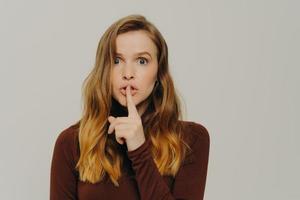Mysterious caucasian teen girl holding finger on lips, looking at camera with scared face expression photo
