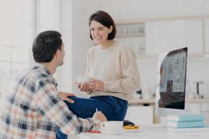 toma interior de una joven feliz y un hombre discuten algo con una bebida, usan una computadora moderna para hacer un informe financiero, posan en una espaciosa sala de luz, colaboran juntos para hacer una tarea común foto