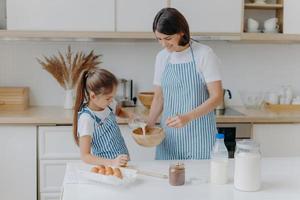 Happy mother and daughter bake together in kitchen, use different ingredients, wear aprons, stand against kitchen interior, girl pours milk in bow. Caring mommy teaches child to cook or make dough