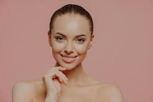 Studio shot of pretty charming woman touches chin, has healthy glowing skin, cares about complexion, stands naked indoor, has cosmetology procedure, isolated on pink background. Beauty concept photo