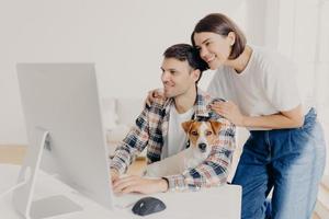 Image of woman touches gently husbands shoulders, look positively in monitor of computer, surf internet, buy furniture online for their new apartment. Happy man works at modern device with dog photo