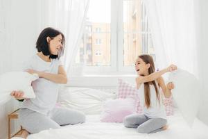 Playful mother and daugher have fun together, have pillow fight on bed, spend free time at home, being in high spirit after sleep, wears casual clothes. Family, togetherness and entertainment photo