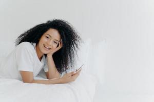 Overjoyed relaxed curly young African American woman enjoys spare time at home, lies in bed with modern cellphone, uses phone in bedroom, sends text message to lover, has lazy morning, good time photo