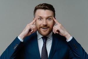 Portrait of handsome male entrepreneur keeps fingers on temples looks with happy surprised expression at camera tries to recall information isolated over grey background. Experienced businessman photo