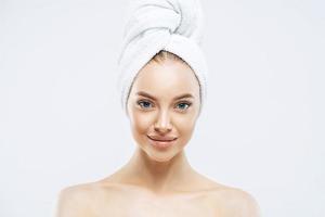 Horizontal shot of charming tender Euoropean woman with healthy skin after taking bath, enjoys hygienic procedures, has natural makeup, wears soft towel on head, stands bare shoulders indoor photo