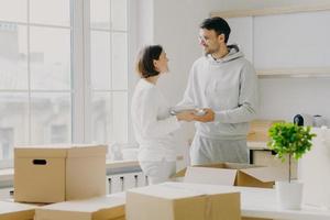 Photo of glad husband and wife carry plates, pose in kitchen near big window, unpack personal belongings from cardboard boxes, dressed in casual outfit, smile pleasantly during talk with each other