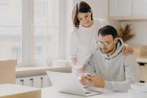 Picture of husband and wife study notification from bank, holds mobile phone and papers, work on laptop computer, pose in kitchen during relocation day, dressed in casual wear, save family money photo