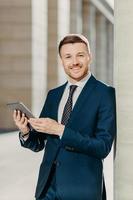 People, work and technology concept. Handsome male economist searches information online on digital tablet, prepares for meeting with staff, works on financial report, dressed in formal clothing photo