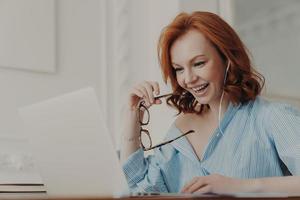 Positive redhead businesswoman participates in online conference, has training class with tutor, focused in display of laptop computer, holds spectacles, enjoys watching educational webinar. photo