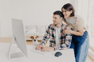 Caring young happy woman leans at husbands shoulders, worries about him, focused into computer monitor, keyboard and surf internet, pose at workplace in modern apartment, little dog sits near photo