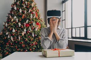 Amazed excited young woman wearing virtual reality goggles sitting against xmas tree