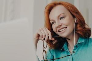 Online studying, communication and distance work concept. Happy professional female freelancer conducts remote course, enjoys e learning in internet, uses earphones, searches information on laptop photo