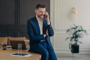 Smiling executive manager talking on phone while sitting on table in office photo