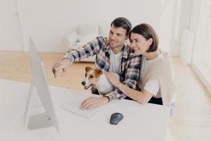 Overjoyed woman and man has happy looks, watch online movie at home together, play with dot, sit at white table, search information via wifi, dressed in casual clothes, Family couple browse internet photo