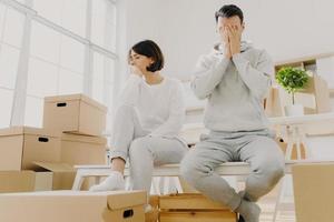 Worried couple pose near stack of cardboard boxes, have to move in other flat, have no money for rent, have dejected sad expressions, sit together in white room, have family conflict and divorce photo