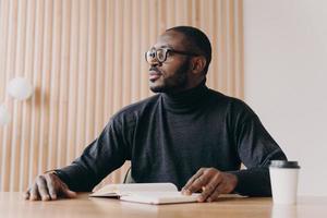 Thoughtful african businessman sits at desk with pen and agenda looks away with pensive expression photo