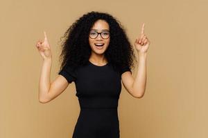 Look upwards. Happy African American woman points above with both index fingers, has slim figure, wears black t shirt, promots object, isolated over brown background. Advertisement and ethnicity photo