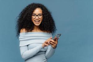 Lovely teenage girl enjoys online communication, holds modern cell phone, checks email box, connected to wireless internet, cannot imagine life without technologies, isolated on blue studio wall photo